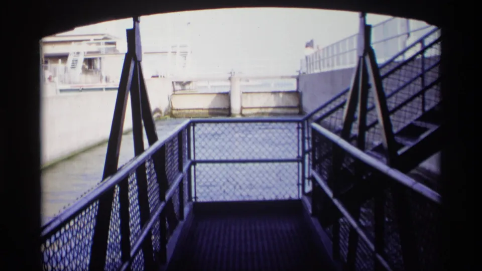 1969 Wisconsin 8mm view from a shadowed metal stair landing toward a concrete canal and pale waterfront structures.