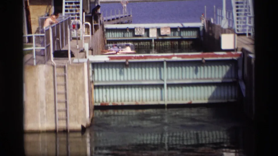 1969 Wisconsin 8mm frame of the lock gate and canal water, with people on the concrete walkway and metal railings above the gate.