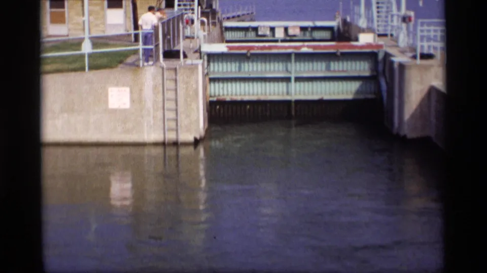 1969 Wisconsin canal lock scene with the greenish metal gate over calm water and people standing along the concrete walkway.