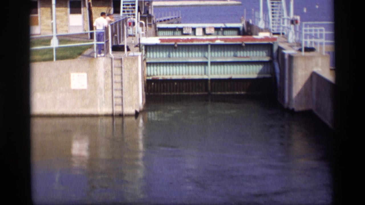 1969 Wisconsin 8mm frame showing a drawbridge gate, railings, water, and two people watching from a concrete walkway.