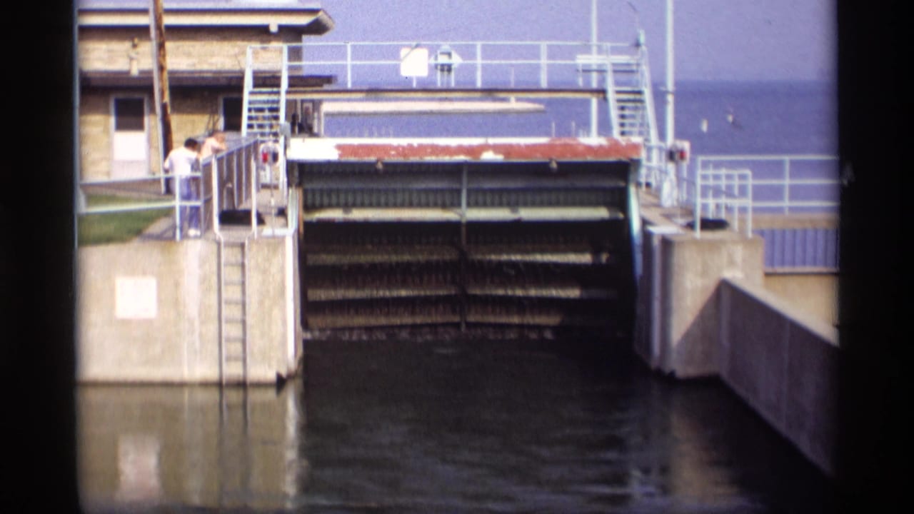 Two people stand on a concrete platform beside a closed slatted sluice gate over dark water.
