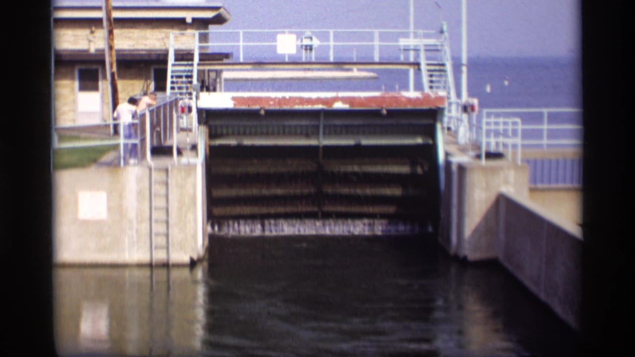 The sluice gate begins opening, with water visible below and two people watching from the side platform.