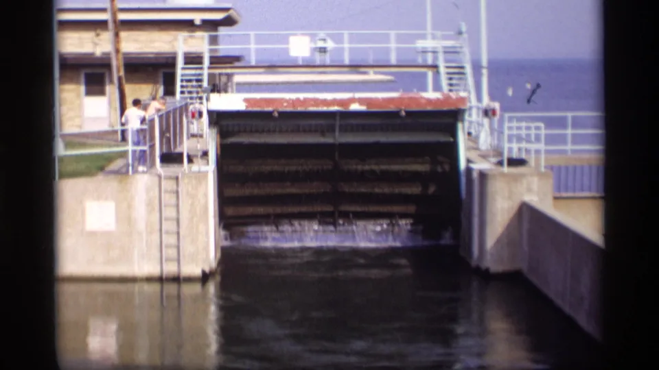 1969 Wisconsin 8mm frame showing water released beneath a raised sluice gate, with two people watching from a concrete platform.