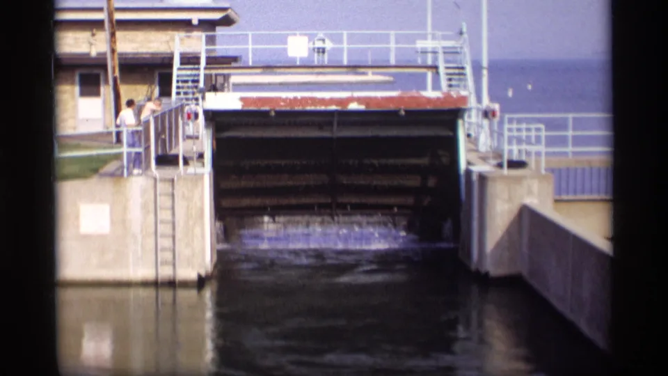 1969 Wisconsin 8mm frame of a raised sluice gate releasing water into a concrete channel while two observers watch nearby.