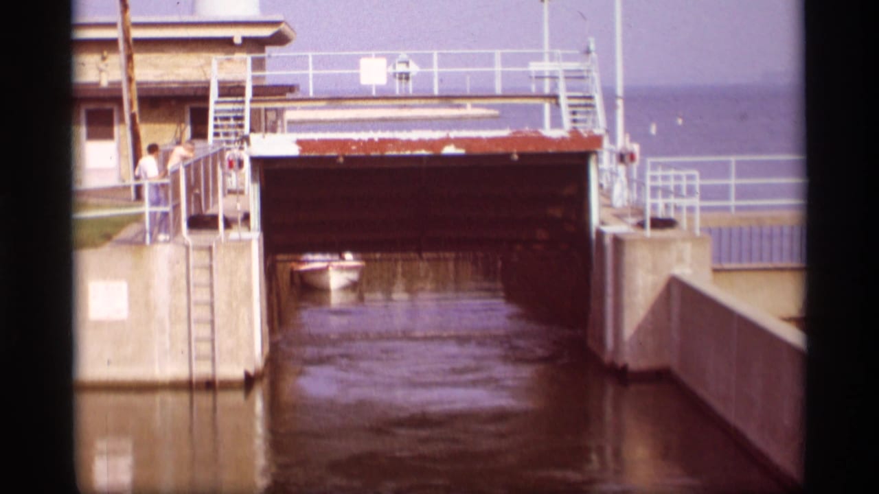 Small boat inside a canal lock beneath a raised gate, with railings and concrete walls around it.