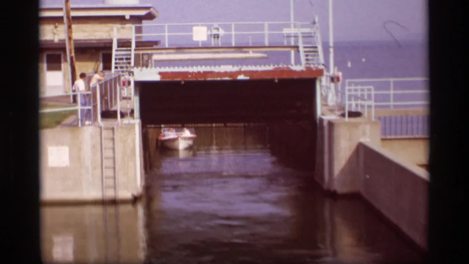 1969 Wisconsin 8mm frame showing a small boat in a canal lock channel, with two people on the left platform and railings above the gate.