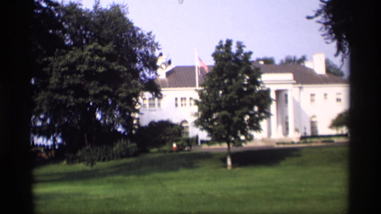 White columned mansion across a green lawn, partly framed by trees and dark film edges.