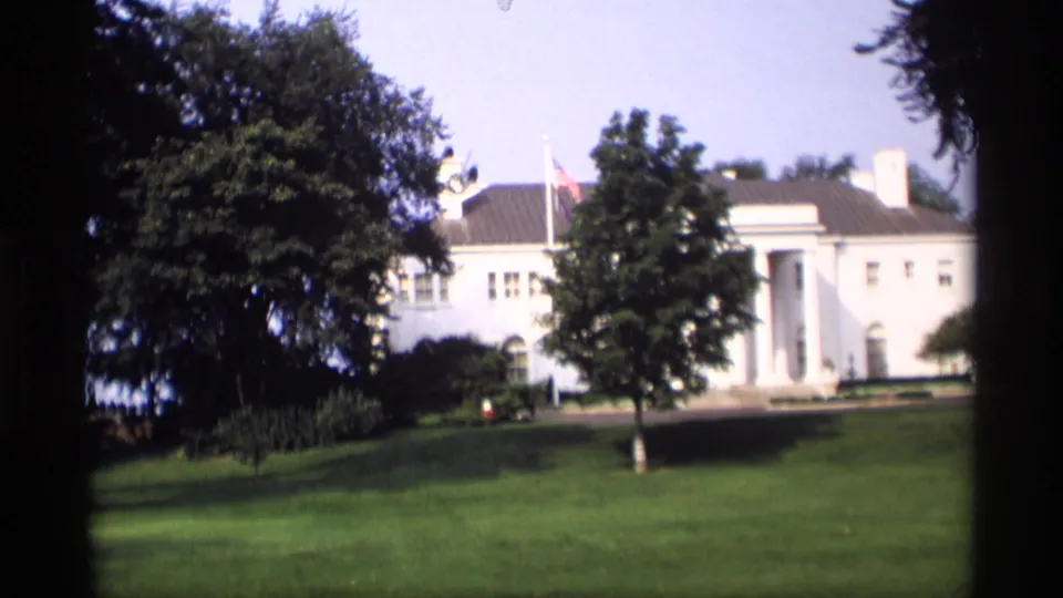 1969 Wisconsin 8mm frame showing a white columned mansion beyond a green lawn, with mature trees and a small flag visible near the roofline.
