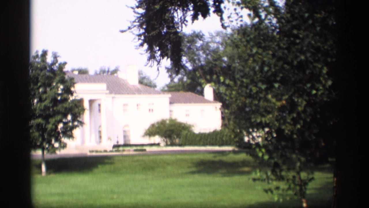 White mansion exterior with columns, lawn, trees, and a dark vertical frame edge.
