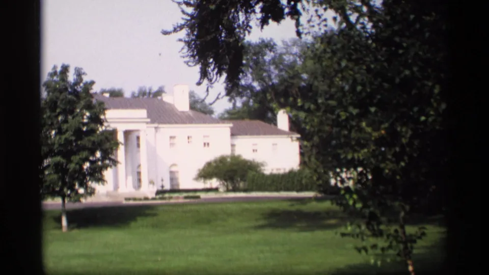 1969 Wisconsin 8mm frame showing the white mansion shifted left in view, with columns, clipped grass, shrubs, and heavy tree cover.