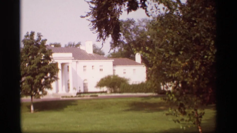 1969 Wisconsin 8mm frame with a warmer exposure shift, showing the white mansion, columned entry, lawn, shrubs, and trees.