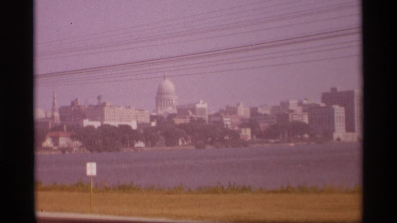 Distant capitol dome across water with skyline, utility lines, grass, and roadside sign.