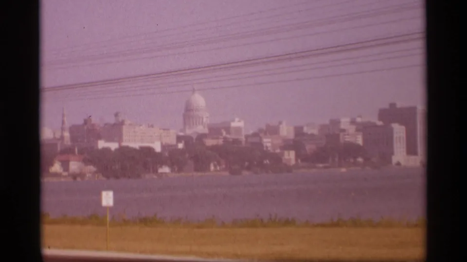 1969 Wisconsin home-movie view of a domed capitol building across water, with skyline buildings, grass verge, utility lines, and a small sign.