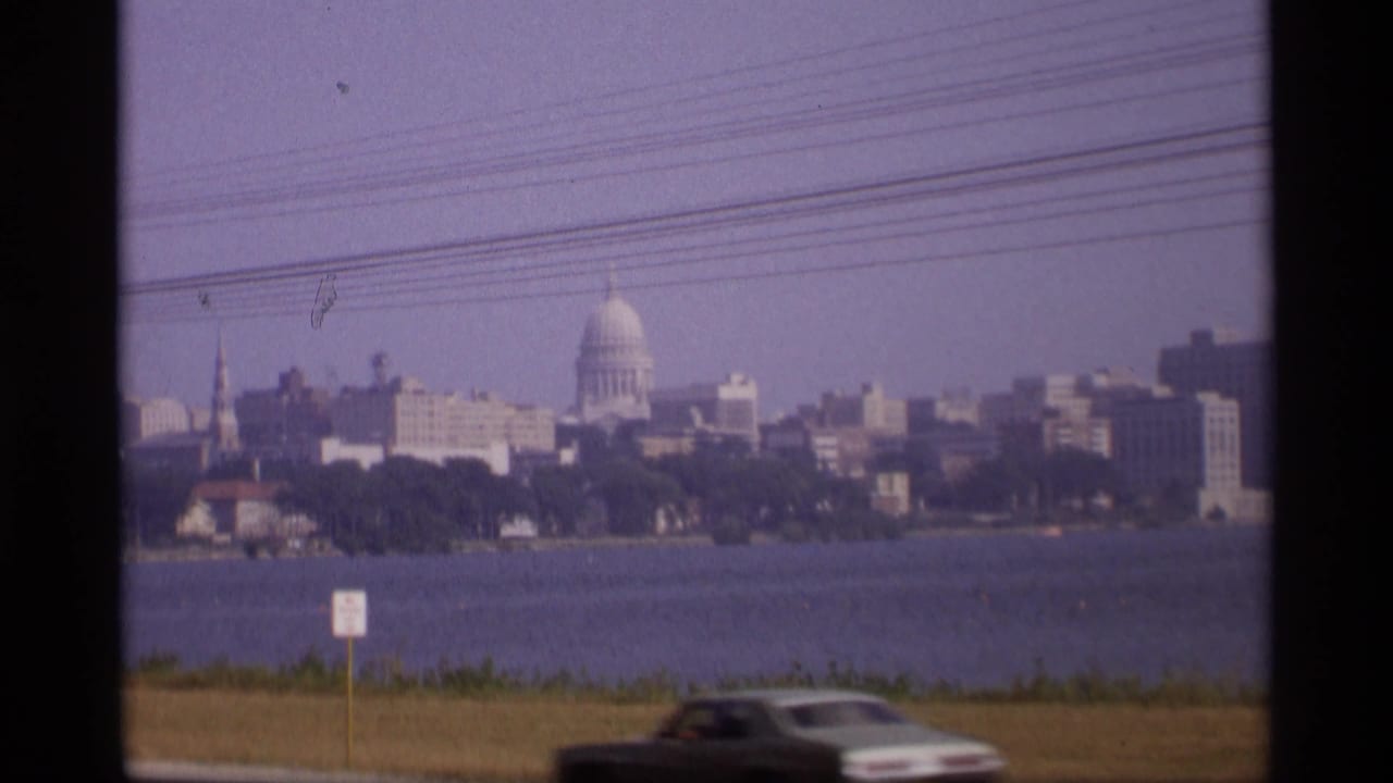1969 Wisconsin view of the capitol dome and waterfront skyline, with overhead lines and a dark car moving through the foreground.