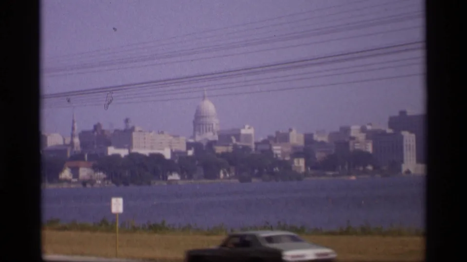 1969 Wisconsin view of the capitol dome and waterfront skyline, with overhead lines and a dark car moving through the foreground.