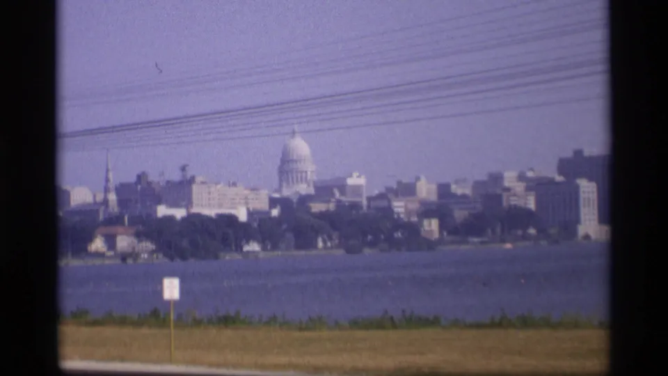 1969 Wisconsin home-movie frame showing the capitol dome across the water, with urban buildings, utility lines, grass, and a roadside sign.