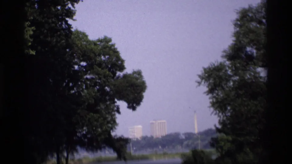 1969 Wisconsin home-movie view framed by trees, looking across water toward distant buildings and a slender vertical landmark or structure.