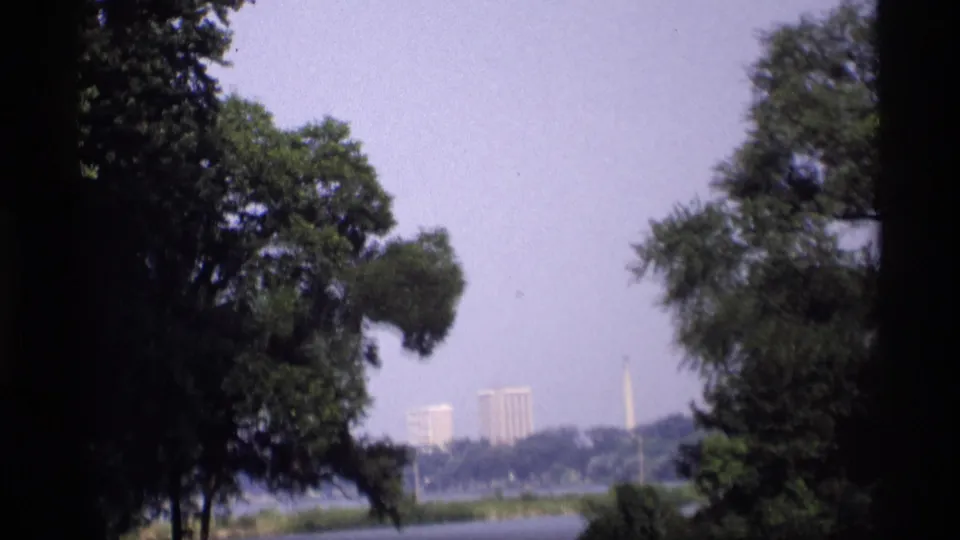 1969 Wisconsin 8mm frame showing a hazy lake view through trees, with distant buildings and a tall monument on the far shore.