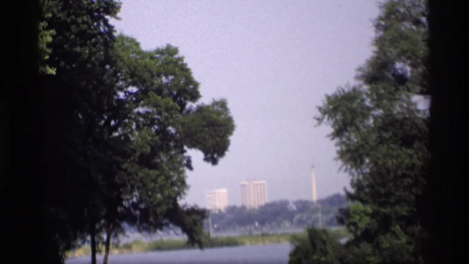 Hazy 1969 Wisconsin lake vista with trees in the foreground and distant buildings and a monument visible across the water.
