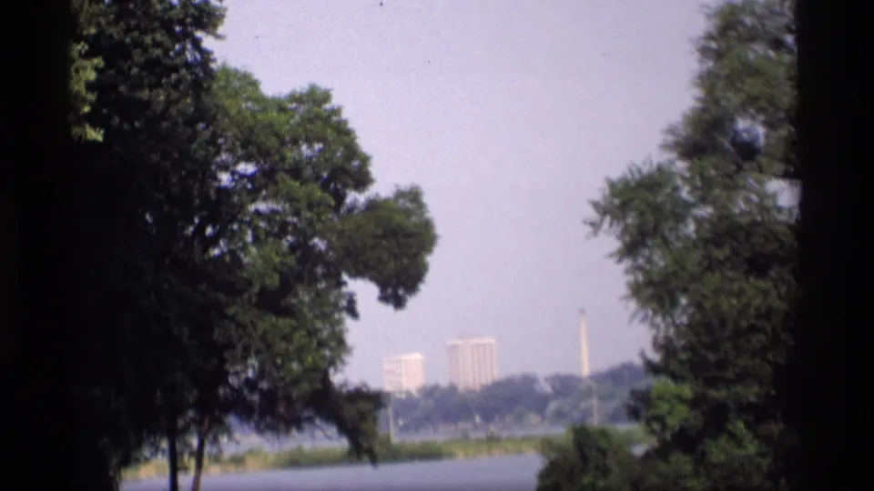 1969 Wisconsin home-movie frame holding on trees, lake water, distant high-rise buildings, and a slender monument in haze.