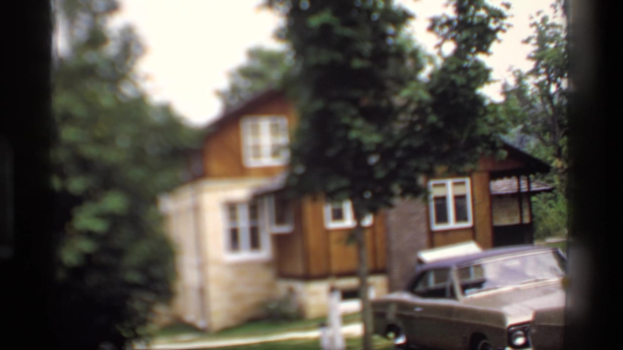 1969 Wisconsin 8mm frame showing a wooded house exterior with stone and wood siding and a period sedan near the driveway.