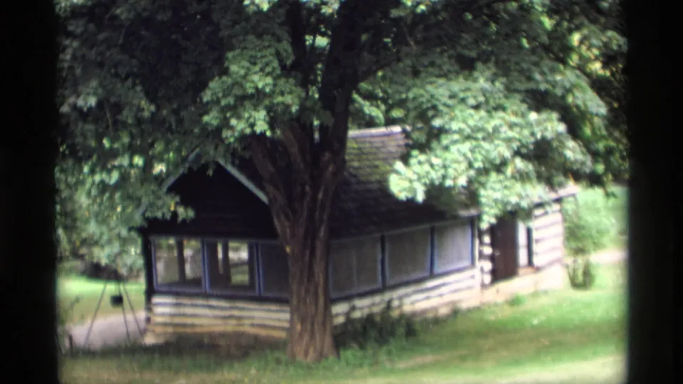1969 Wisconsin 8mm frame showing a small rustic cabin or outbuilding beneath dense trees in a wooded yard.