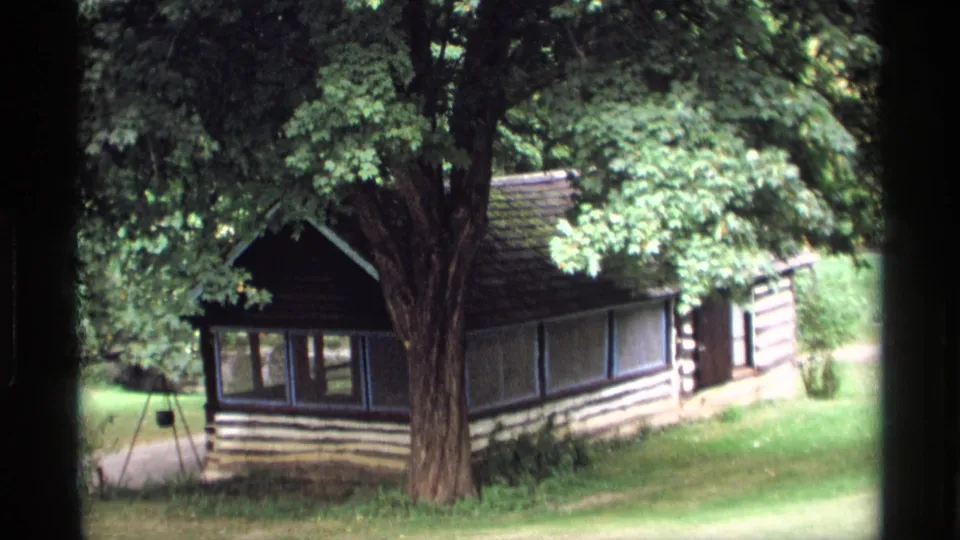 1969 Wisconsin home-movie still of a small cabin-like structure shaded by a broad tree in a quiet wooded yard.