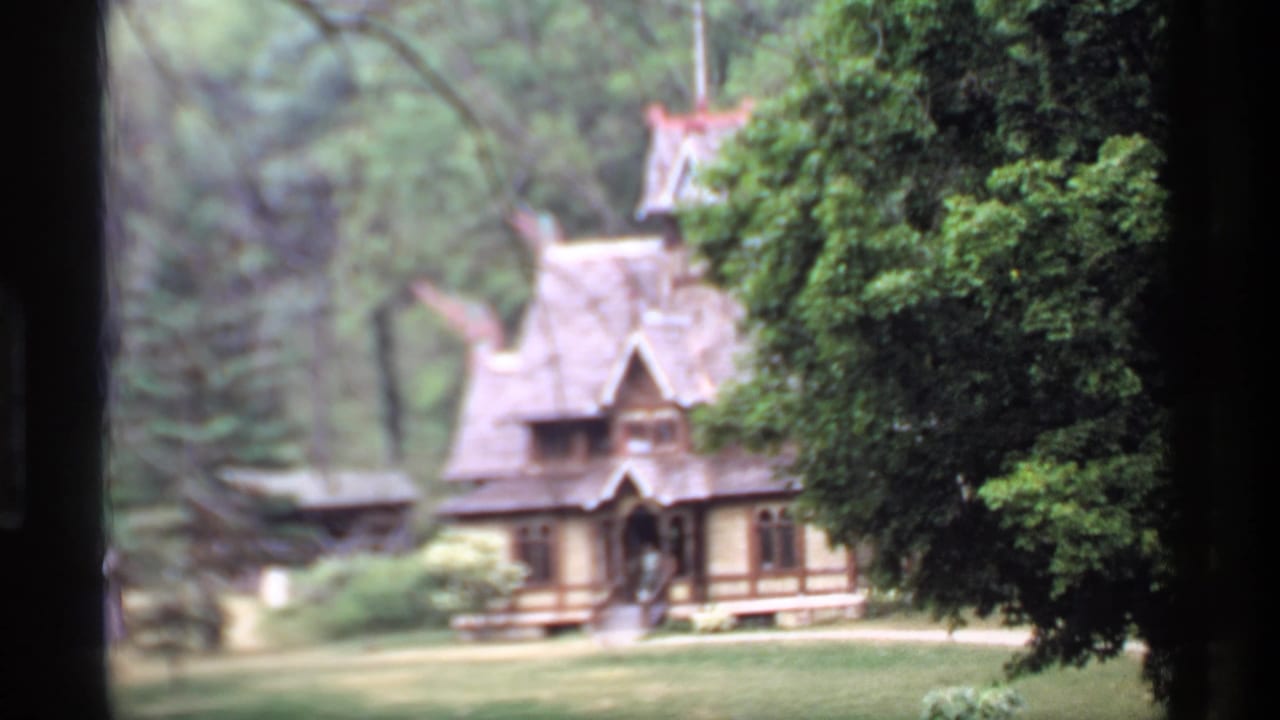 Ornate wooden house with steep roof seen through dark foreground framing and trees.