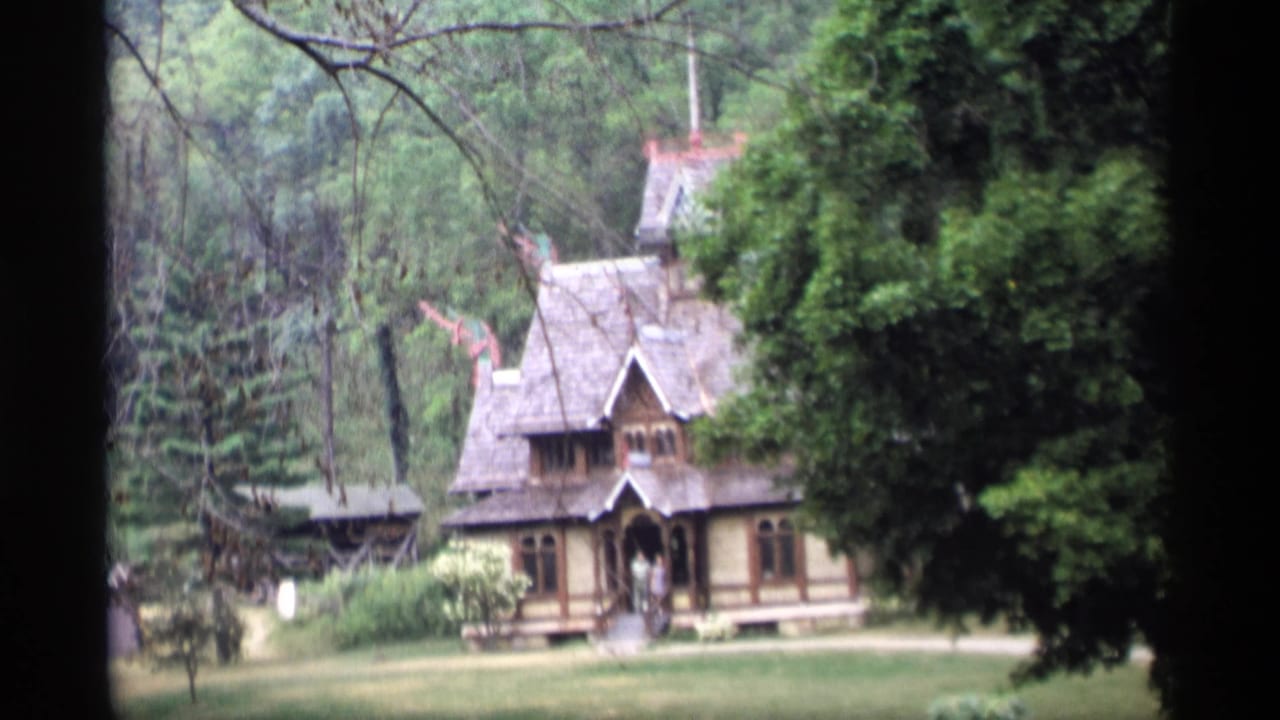 Rustic house with steep gables and surrounding trees viewed from a shaded opening.