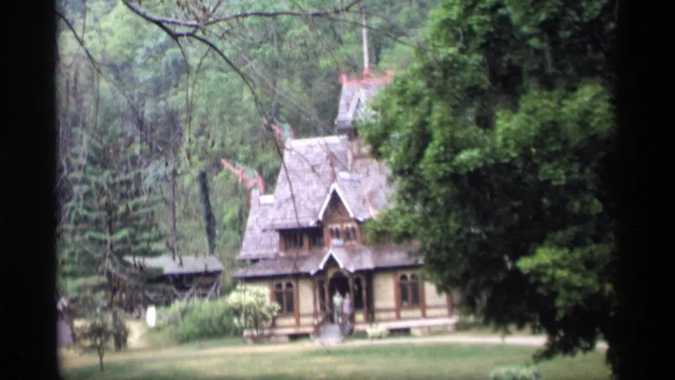 1969 Wisconsin frame showing a decorative rustic house on a lawn, with dense trees and a small person near the entry.