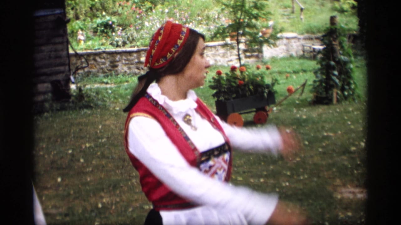 Woman in red and white folk costume gestures outdoors beside garden flowers.