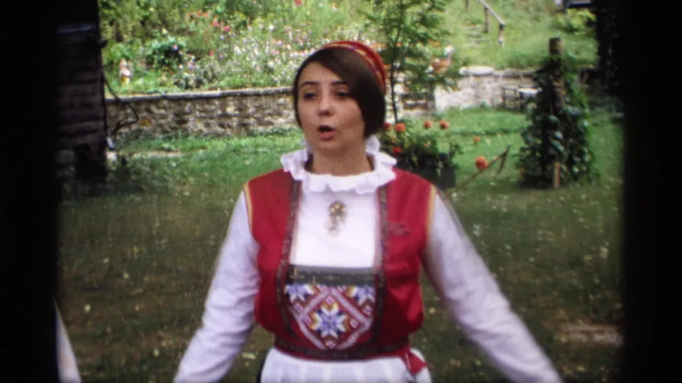 1969 Wisconsin 8mm frame showing a woman in folk-style dress facing the camera in a green garden setting.