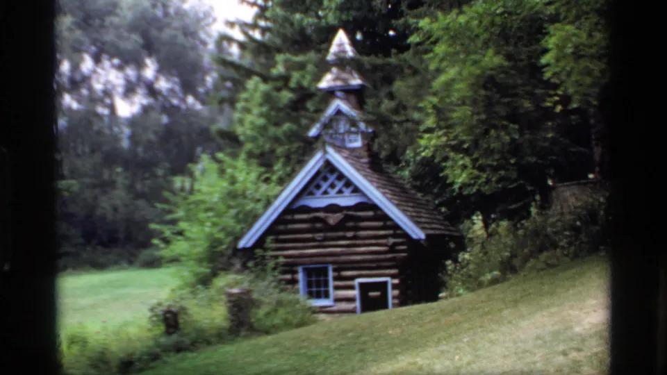 1969 Wisconsin 8mm frame of a small log cabin-like building with a steep roof set in a wooded grassy area.