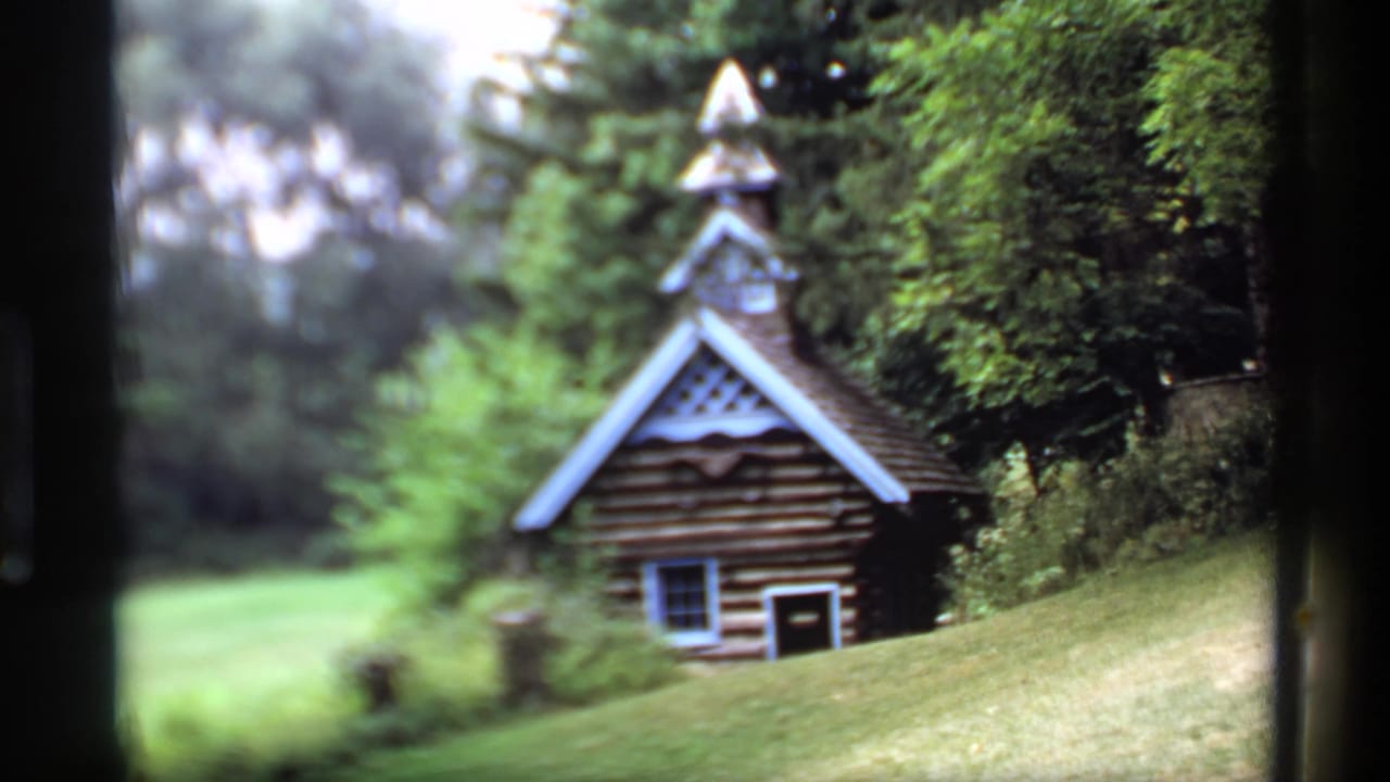 Small log chapel-like building with a pointed roof beside grass and trees.