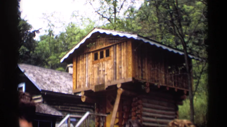 1969 Wisconsin home-movie frame of a raised wooden room, log walls, shingled roof, and wooded background.