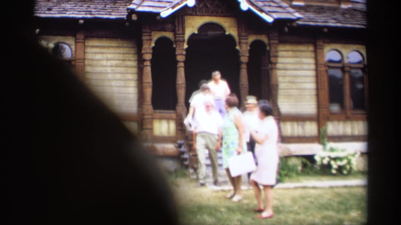 Older adults stand near the entrance of an ornate wooden house with arched porch trim.