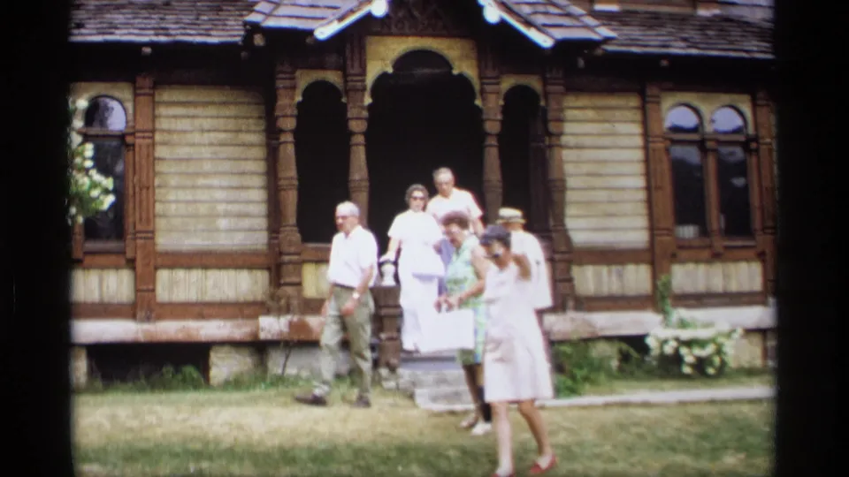 1969 Wisconsin home-movie frame showing older adults leaving or socializing on a lawn before an ornate wooden house.