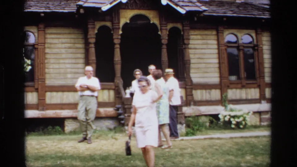 A woman in a pale dress walks toward the camera as older adults linger outside the ornate wooden house in 1969 Wisconsin footage.