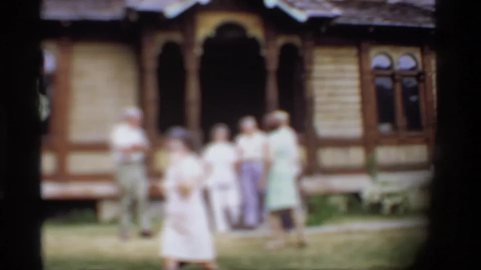 Soft 1969 Wisconsin home-movie frame showing adults on a lawn before a historic-looking wooden building with arched trim.