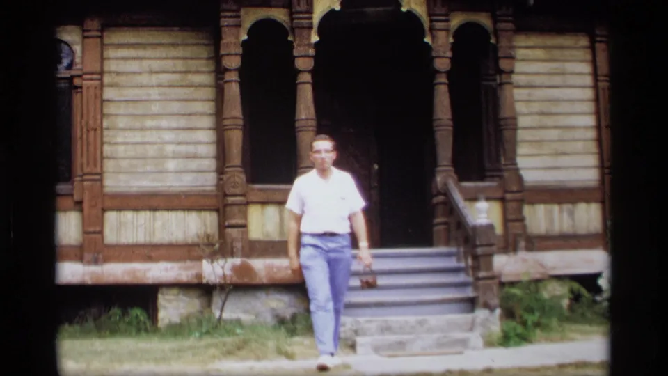 1969 Wisconsin 8mm frame of a man walking from the steps of an older house with arched porch openings and wooden trim.