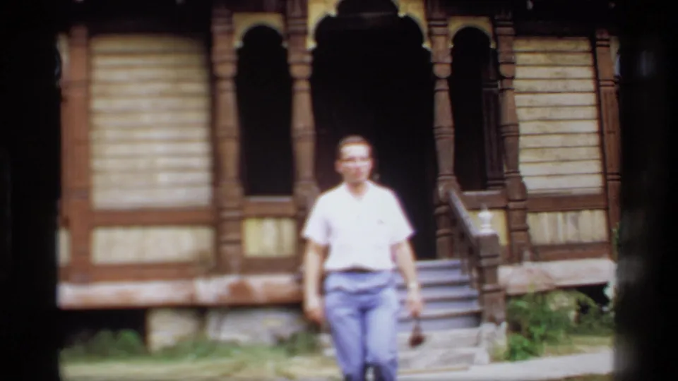 Softly focused 1969 Wisconsin home-movie frame of a man walking past the steps and porch of an older wooden house.