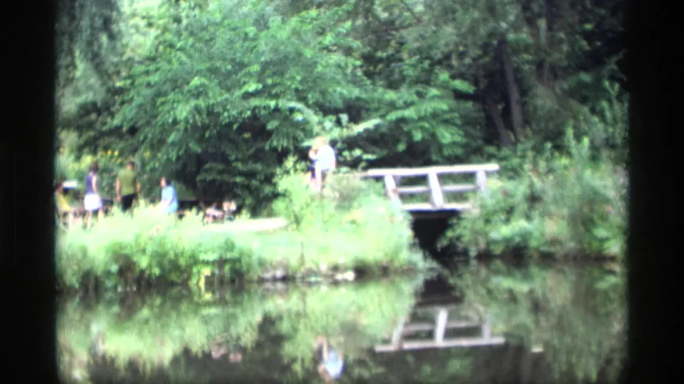 1969 Wisconsin home-movie frame of visitors by a pond and small wooden footbridge, with trees and bridge reflections in the water.