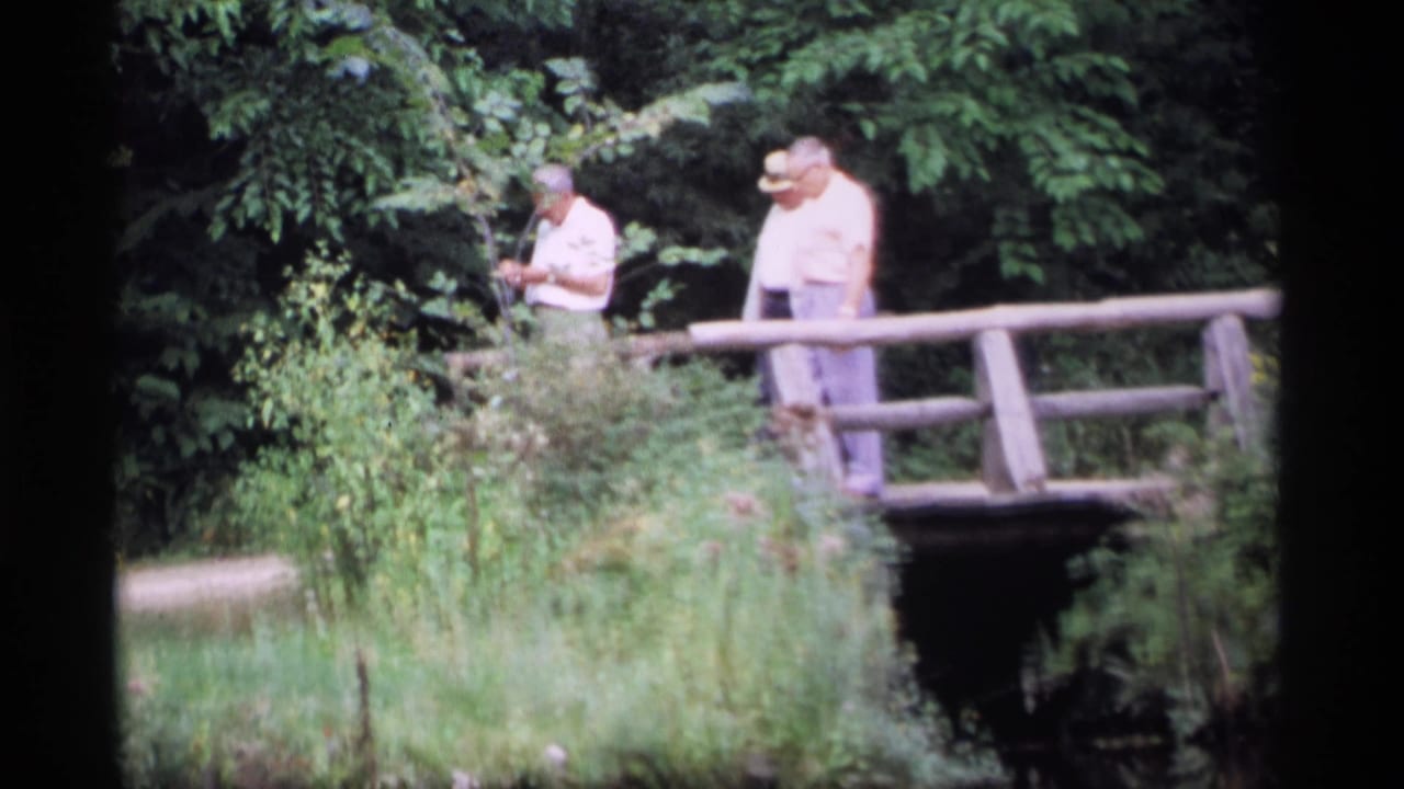 Three older men walk or stand on a rustic wooden bridge in a leafy wooded area.