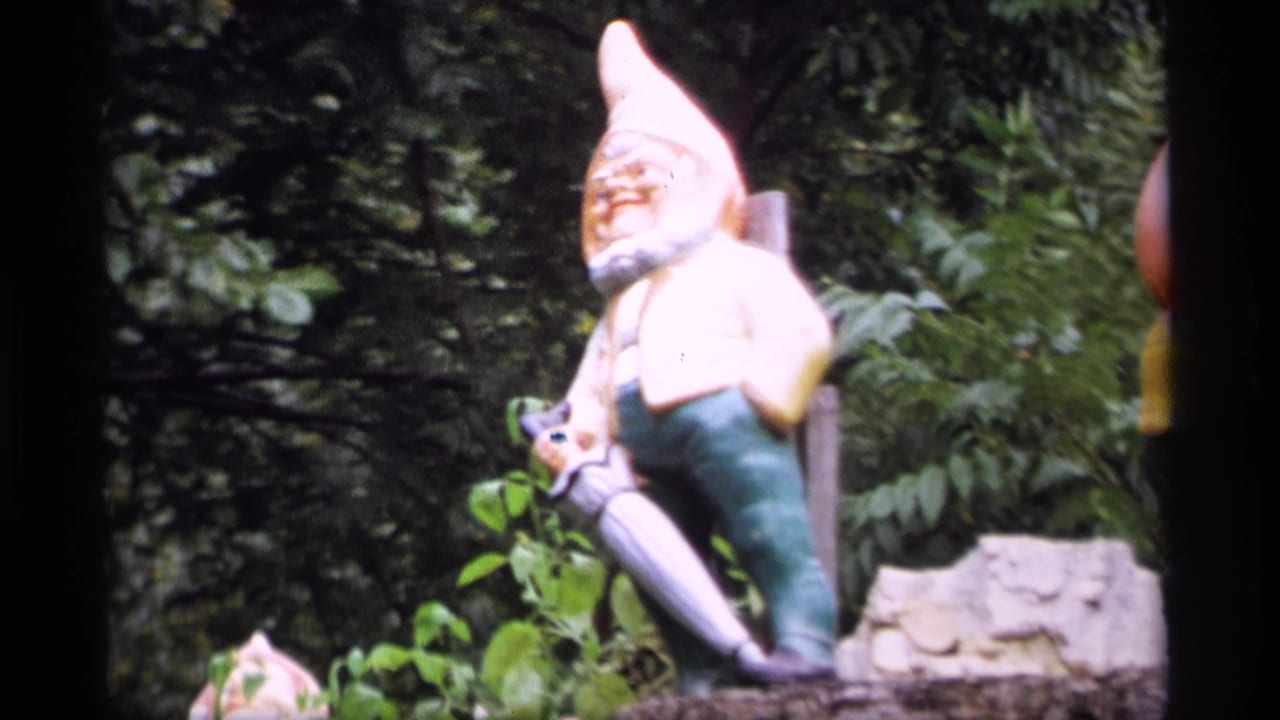 1969 Wisconsin 8mm frame of a garden gnome statue with a tall pale hat, green pants, and shovel amid dense foliage.