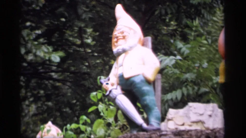 1969 Wisconsin 8mm frame of a garden gnome statue with a tall pale hat, green pants, and shovel amid dense foliage.