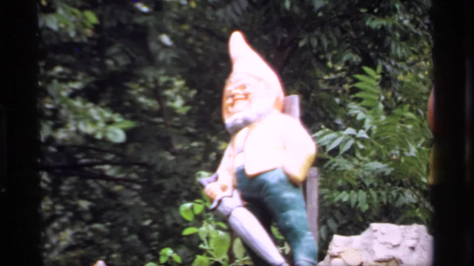 Soft 1969 Wisconsin home-movie view of a painted garden gnome figure standing near shrubs and low stonework.
