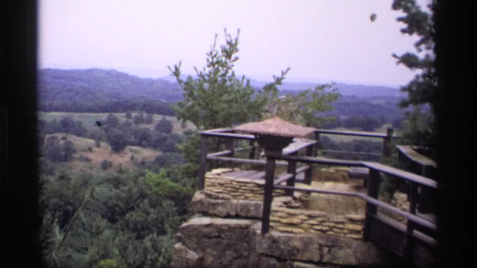 Vintage 1969 Wisconsin 8mm frame with a wooden-railed stone lookout in front of a broad wooded landscape.