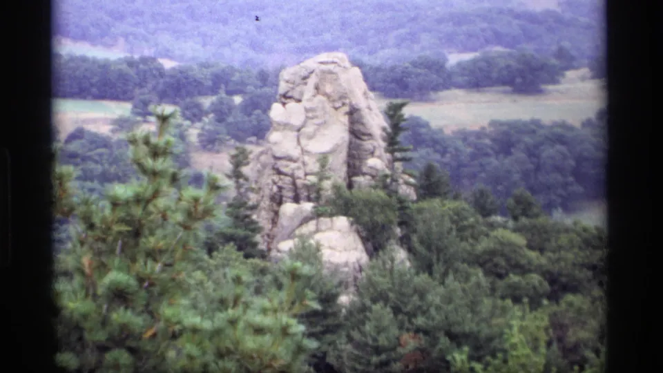 1969 Wisconsin 8mm frame showing a pale rock spire above dense green forest and rolling hills under a hazy sky.