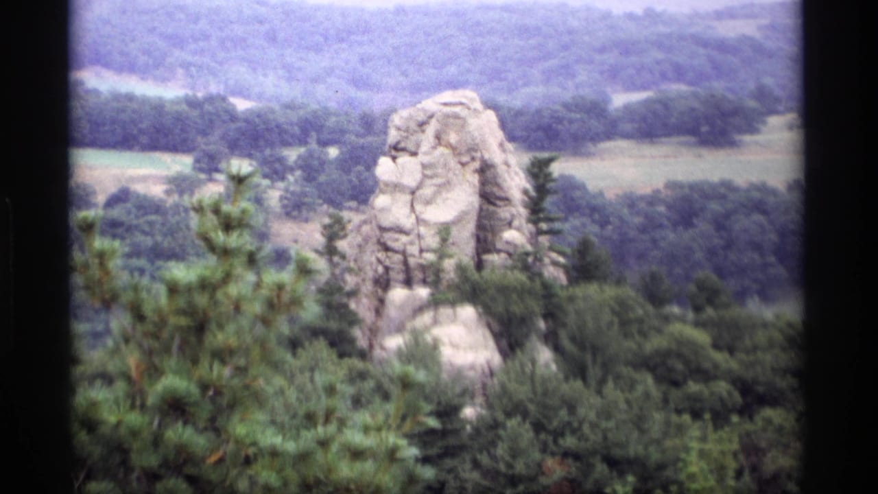 Rock formation centered beyond trees, with wooded hills and fields in the distance.