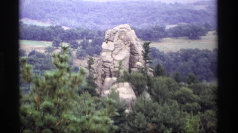 1969 Wisconsin 8mm view of a natural stone formation surrounded by trees, with distant rural fields beyond.